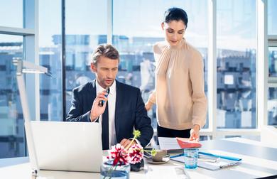 A man in a business suit sits at an office desk with a laptop, holding a voice recorder while a woman standing next to him hands over a red folder